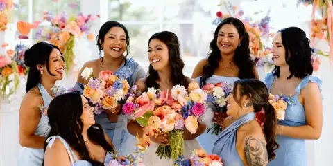 Bride and bridesmaids in light blue dresses smiling with colorful bouquets at a wedding.