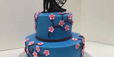 A two-tiered blue wedding cake decorated with pink sugar flowers and a black Mr and Mrs silhouette topper.