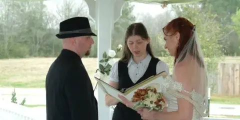 A woman officiating a wedding ceremony for a bride and groom in a white gazebo.