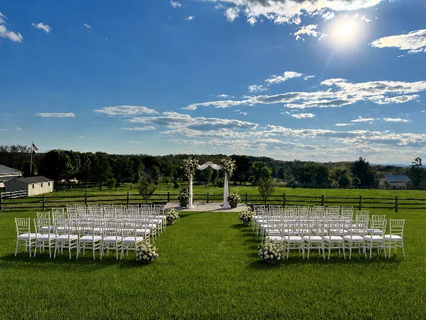 An outdoor wedding ceremony setup on a grassy lawn featuring a floral arch and rows of white chairs under a blue sky.