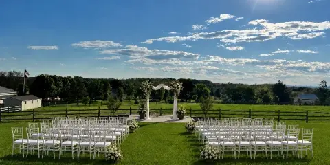 An outdoor wedding ceremony setup on a grassy lawn featuring a floral arch and rows of white chairs under a blue sky.
