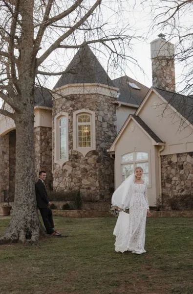 A bride in a white lace gown and a groom in a dark suit standing on a lawn in front of a stone and stucco house.