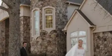 A bride in a white lace gown and a groom in a dark suit standing on a lawn in front of a stone and stucco house.