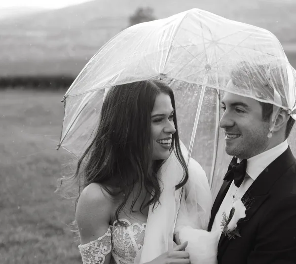 A bride and groom smiling at each other under a clear umbrella in a field.