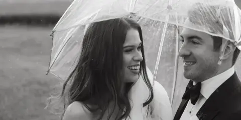 A bride and groom smiling at each other under a clear umbrella in a field.