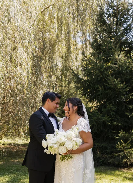 A bride and groom standing outdoors, with the bride holding a large white floral bouquet.