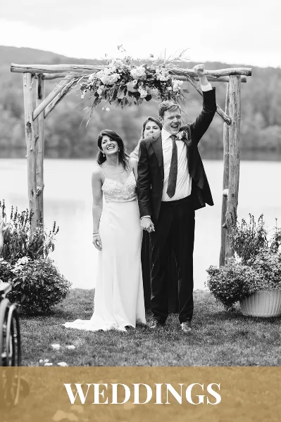 A black and white photograph of a bride and groom standing under a rustic wooden arch decorated with flowers, with a body of water and hills in the background.