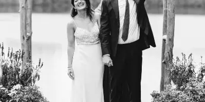 A black and white photograph of a bride and groom standing under a rustic wooden arch decorated with flowers, with a body of water and hills in the background.