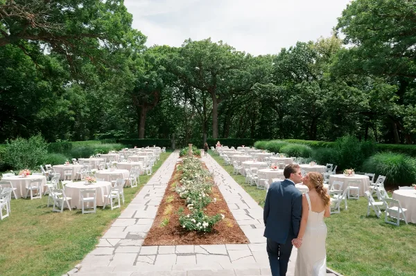 Outdoor wedding reception setup with rows of round tables covered in white linens and white folding chairs arranged in a garden setting.