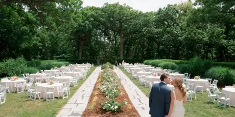 Outdoor wedding reception setup with rows of round tables covered in white linens and white folding chairs arranged in a garden setting.