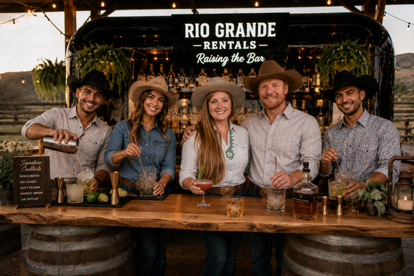 A group of five professional bartenders in western attire standing behind a rustic wooden bar with the 'Rio Grande Rentals' sign, serving drinks.