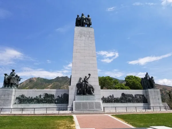 The 'This Is The Place' monument featuring a tall stone obelisk with bronze statues of pioneers at the top and base, set against a blue sky and mountains.