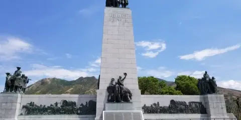 The 'This Is The Place' monument featuring a tall stone obelisk with bronze statues of pioneers at the top and base, set against a blue sky and mountains.