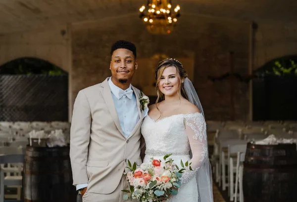 A bride and groom standing in a rustic brick-walled venue with a chandelier and wooden barrels.