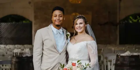 A bride and groom standing in a rustic brick-walled venue with a chandelier and wooden barrels.