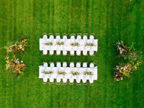 Aerial view of a luxury outdoor wedding reception setup with two long tables, floral centerpieces, and gold chairs on a green lawn.