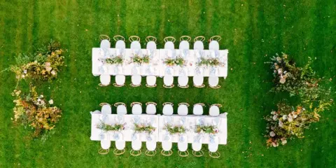 Aerial view of a luxury outdoor wedding reception setup with two long tables, floral centerpieces, and gold chairs on a green lawn.