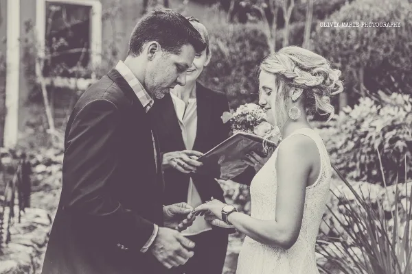 A black and white photograph of a bride and groom exchanging rings during an outdoor wedding ceremony, with an officiant standing between them.
