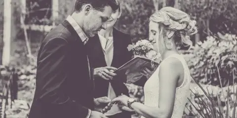 A black and white photograph of a bride and groom exchanging rings during an outdoor wedding ceremony, with an officiant standing between them.