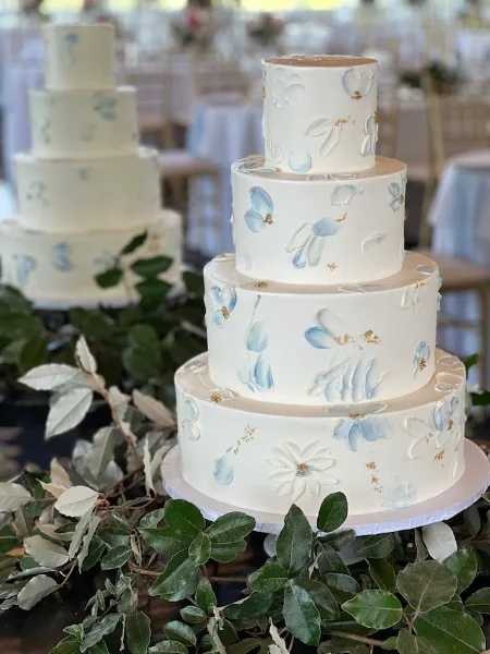 A four-tier white wedding cake decorated with hand-painted blue and white flowers and gold accents, displayed on a stand with greenery.