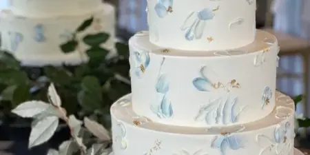 A four-tier white wedding cake decorated with hand-painted blue and white flowers and gold accents, displayed on a stand with greenery.
