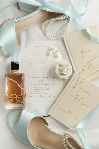 A flat-lay photograph of wedding invitations, rings, perfume, and accessories arranged on a white background.