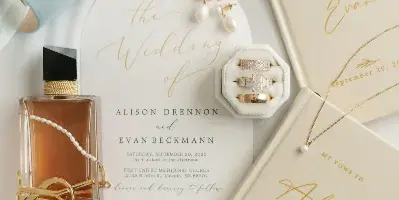 A flat-lay photograph of wedding invitations, rings, perfume, and accessories arranged on a white background.