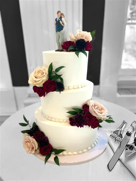 A three-tiered white wedding cake decorated with red and cream roses, dark flowers, and a couple figurine topper, sitting on a table with silver cake serving utensils.