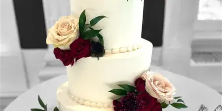A three-tiered white wedding cake decorated with red and cream roses, dark flowers, and a couple figurine topper, sitting on a table with silver cake serving utensils.