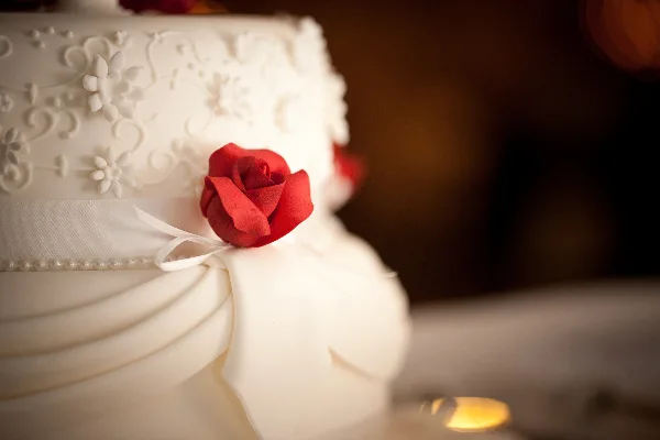 A close-up photograph of a white tiered cake featuring white floral piping, a white ribbon, and a red sugar rose.