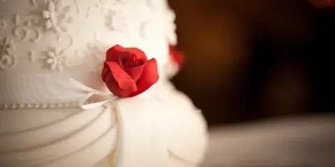 A close-up photograph of a white tiered cake featuring white floral piping, a white ribbon, and a red sugar rose.