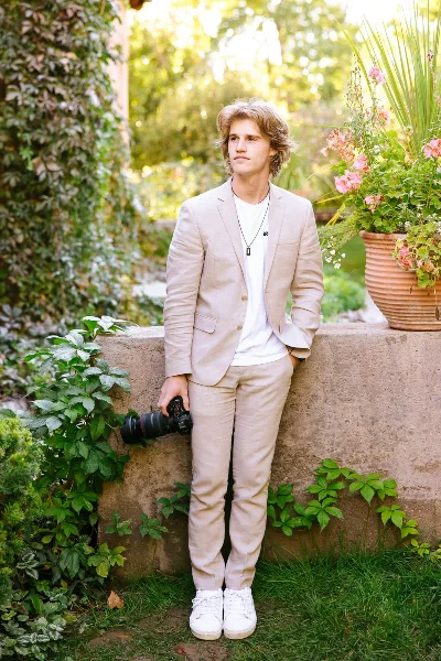 A young man in a light beige suit stands in a garden holding a professional camera with a large lens.