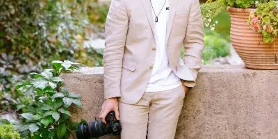 A young man in a light beige suit stands in a garden holding a professional camera with a large lens.