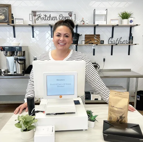 A smiling woman stands behind a counter with a POS system displaying 'Fresh & Fancy @ Mama Sheenas' in a kitchen setting.