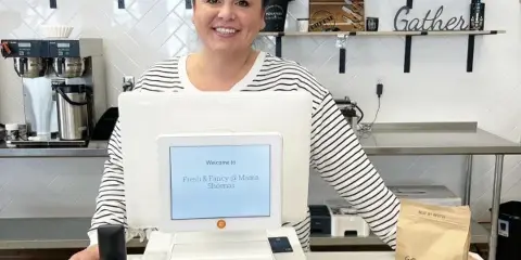 A smiling woman stands behind a counter with a POS system displaying 'Fresh & Fancy @ Mama Sheenas' in a kitchen setting.