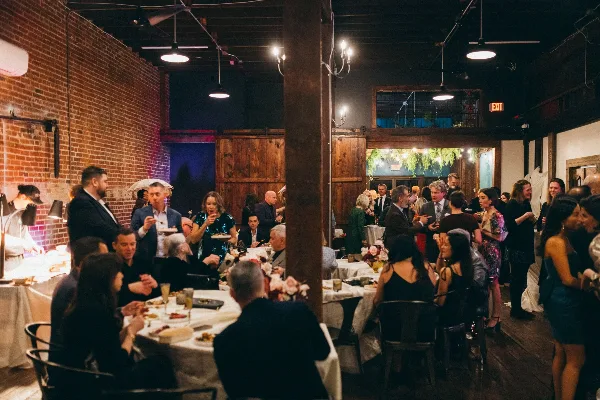 Guests dining and mingling at a wedding reception inside a brick-walled event venue with round tables and warm lighting.
