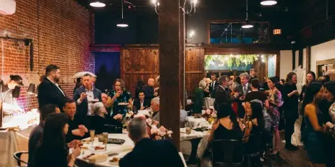 Guests dining and mingling at a wedding reception inside a brick-walled event venue with round tables and warm lighting.