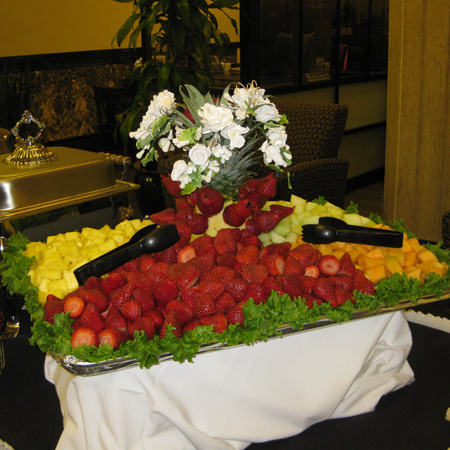 A catering buffet table featuring a large arrangement of fresh strawberries, pineapple, and melon with serving tongs, garnished with lettuce and a white rose centerpiece, set in an event venue.