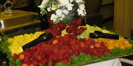 A catering buffet table featuring a large arrangement of fresh strawberries, pineapple, and melon with serving tongs, garnished with lettuce and a white rose centerpiece, set in an event venue.
