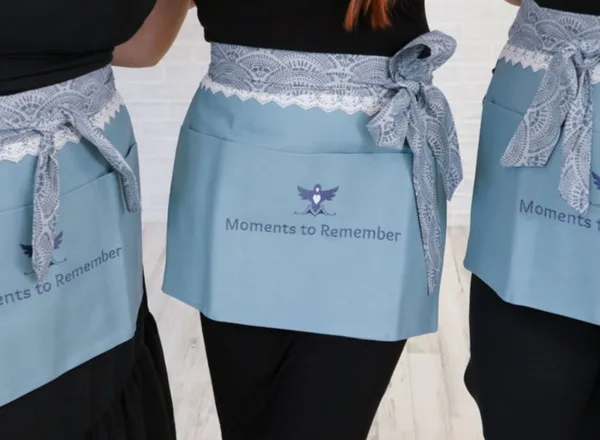 Close-up of light blue aprons with lace trim, featuring the 'Moments to Remember' logo and name.