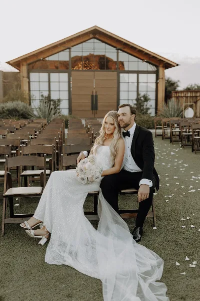 A bride and groom sitting on a chair in front of a wooden venue building with rows of ceremony chairs and desert plants.