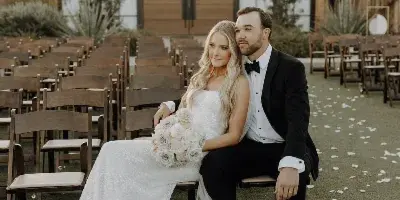 A bride and groom sitting on a chair in front of a wooden venue building with rows of ceremony chairs and desert plants.