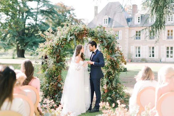 A bride and groom exchanging vows under a floral arch at an outdoor wedding ceremony.