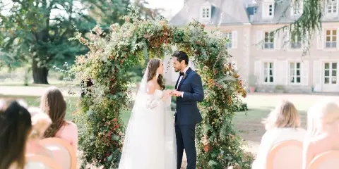 A bride and groom exchanging vows under a floral arch at an outdoor wedding ceremony.