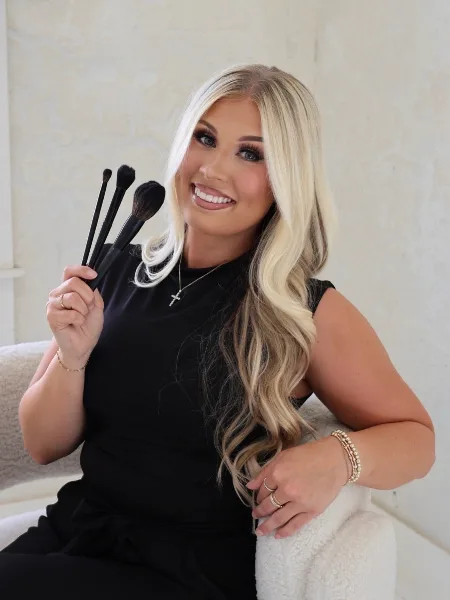 A smiling woman with long blonde hair holding a set of makeup brushes.