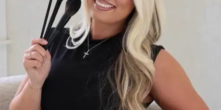 A smiling woman with long blonde hair holding a set of makeup brushes.