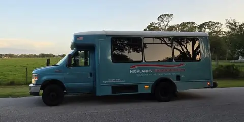 A teal shuttle bus with 'Highlands Transportation' branding parked on a road near a grassy field.