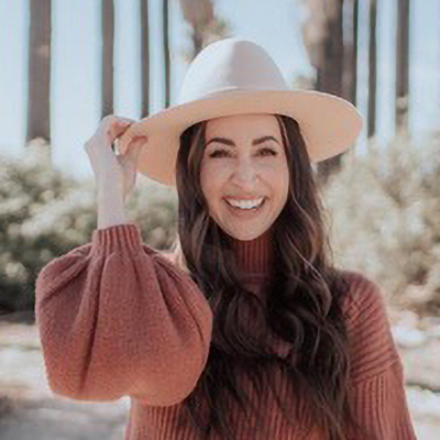 Portrait of a smiling woman with long dark hair wearing a wide-brimmed hat and rust-colored sweater outdoors.