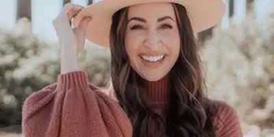 Portrait of a smiling woman with long dark hair wearing a wide-brimmed hat and rust-colored sweater outdoors.