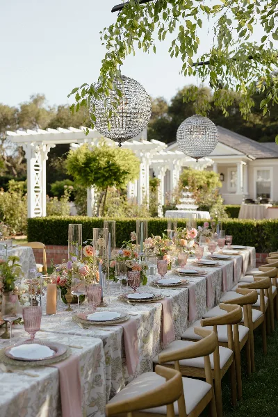 Outdoor wedding reception table set with floral arrangements, glassware, and hanging crystal orbs under a leafy canopy.